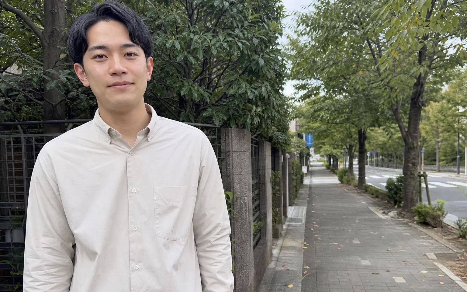 曇りの日の並木道で、無地のシャツを着て街路樹の前に立つ男性